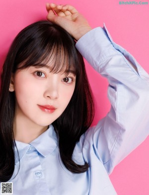 A woman in a blue shirt and gray skirt sitting on a pink background.